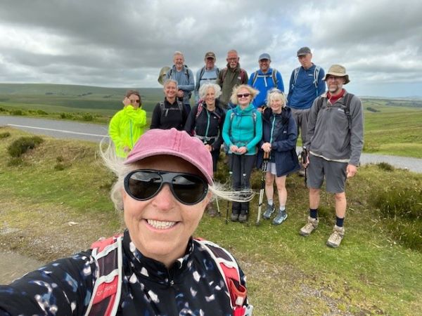 Woman in foreground wearing hat and sunglasses with group of walkers behind on grassy ground with sky with grey clouds